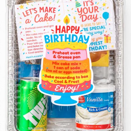 Finished birthday cake donation kit assembled in a rectangular 9×13-inch aluminum pan with a clear lid, colorful cake-shaped birthday label, baking instructions, frosting, soda, and birthday card.