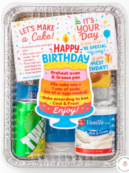 Finished birthday cake donation kit assembled in a rectangular 9×13-inch aluminum pan with a clear lid, colorful cake-shaped birthday label, baking instructions, frosting, soda, and birthday card.