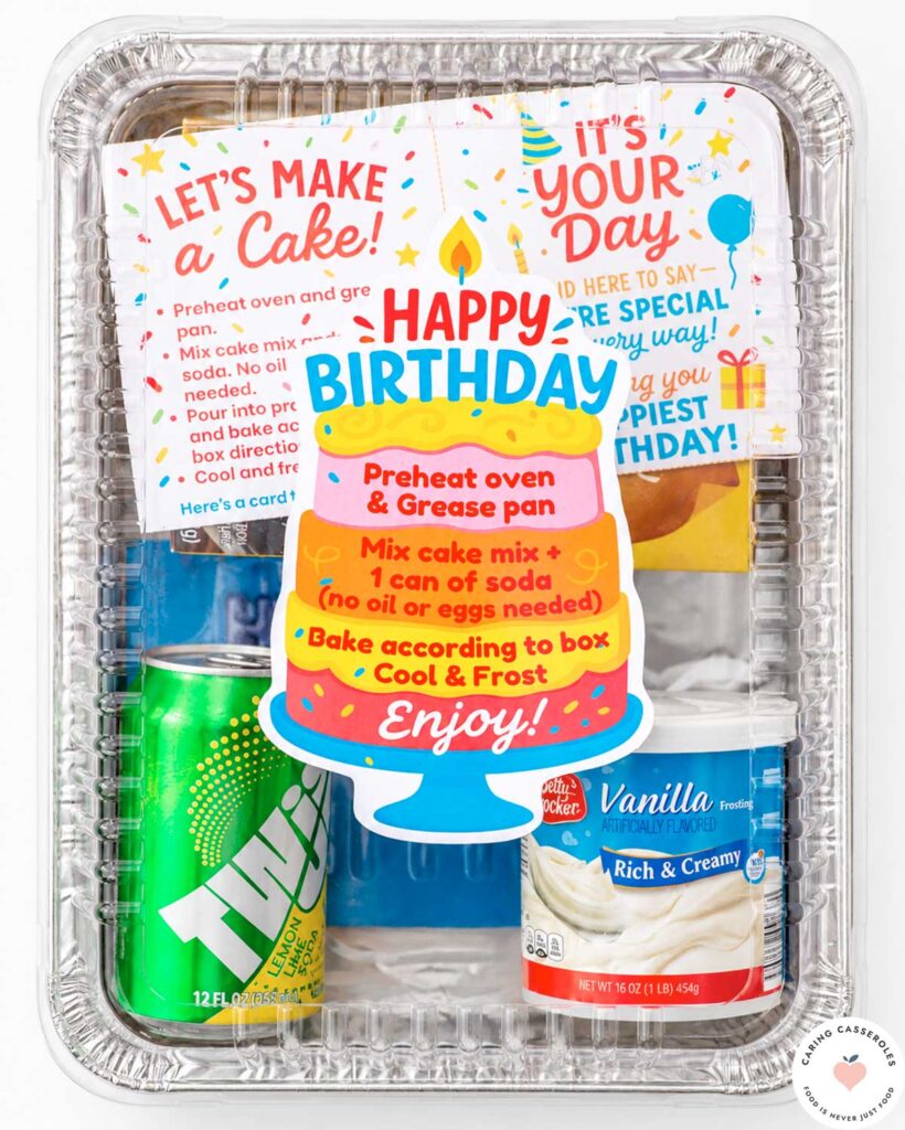 Finished birthday cake donation kit assembled in a rectangular 9×13-inch aluminum pan with a clear lid, colorful cake-shaped birthday label, baking instructions, frosting, soda, and birthday card.
